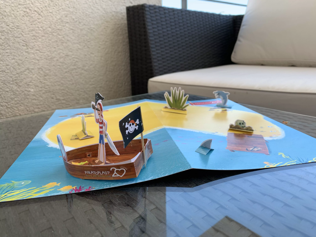 Nahaufnahme von Bastelbogen: Pirateninsel, Piratenschiff mit Piratoplast-Flagge, Piratenjungen und Haifischflosse im Wasser