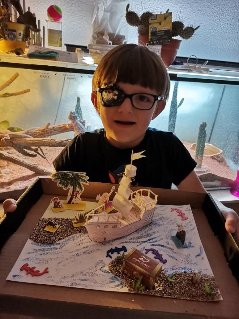 Junge mit Piratoplast Augenpflaster zeigt ein gebasteltes Piratenschiff und Elemente des Bastelbogens in die Kamera