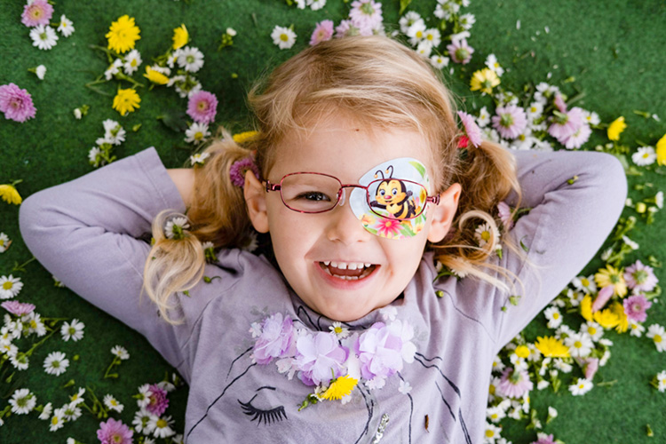 Mädchen mit Augenpflaster und Brille liegt auf einer Blumenwiese und lacht in die Kamera