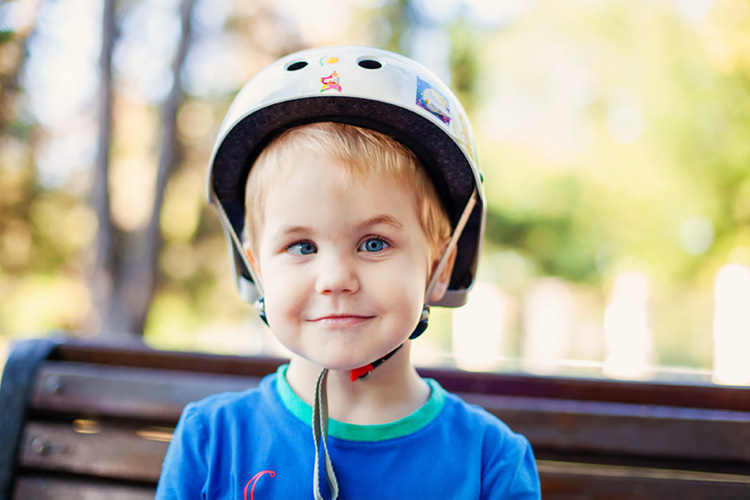 Junge mit Strabismus sitzt auf einer Parkbank mit einem Fahrradhelm auf dem Kopf und lächelt in die Kamera