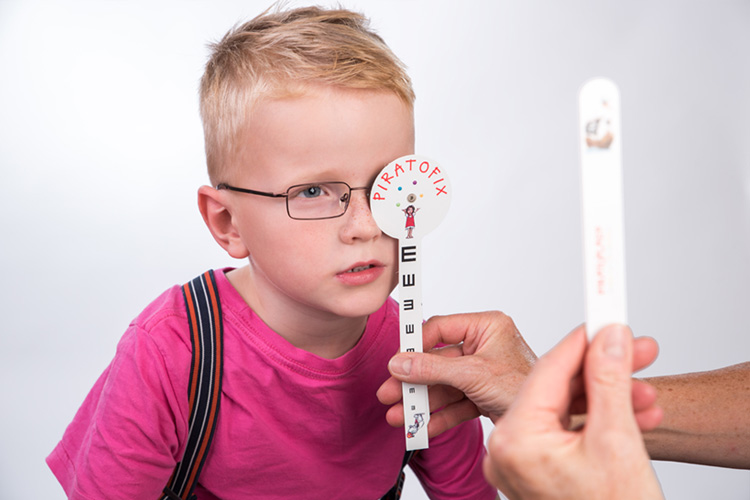 Kleiner Junge mit Brille macht einen Sehtest: Ein Auge wird verdeckt und er fokussiert eine Testkarte